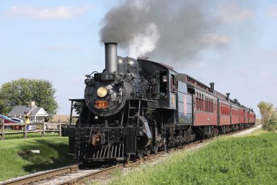 A vintage American steam locomotive hauls a rake of even more vintage bogie passenger coaches along a dead straight track through a flat landscape on a heritage railway in the USA.